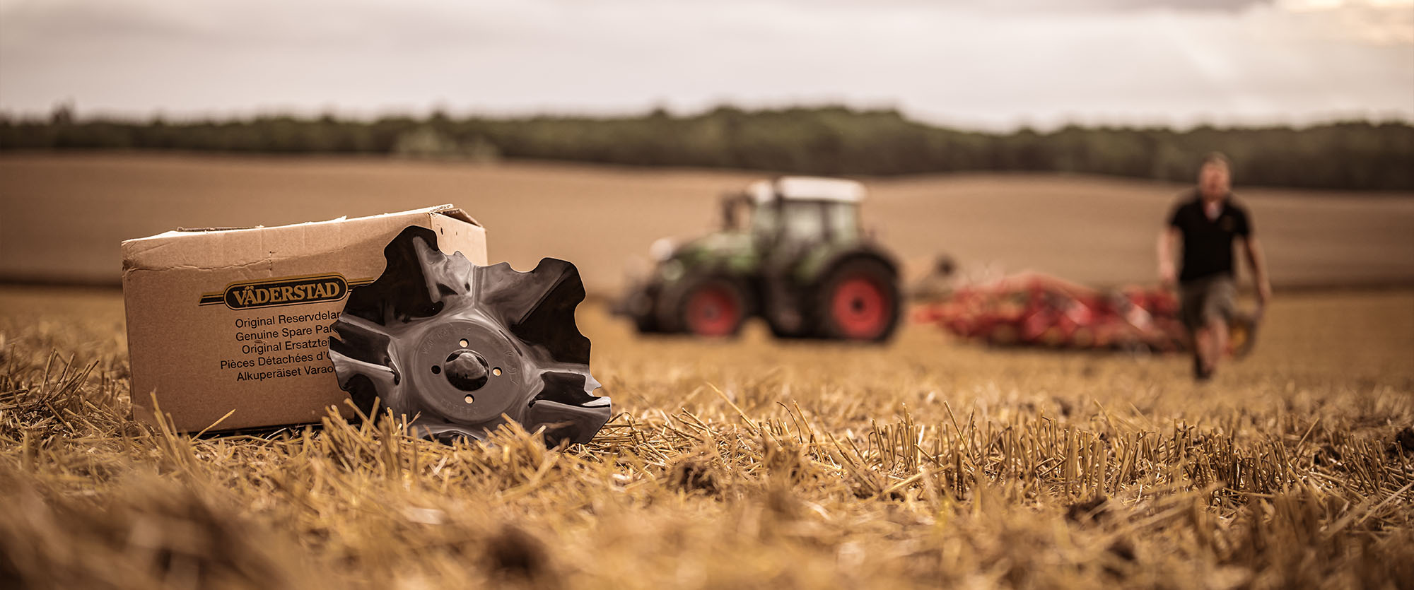A Väderstad cultivator disc is leaning against a box in a field. In the background is a tractor and a cultivator, the farmer is walking towards us.